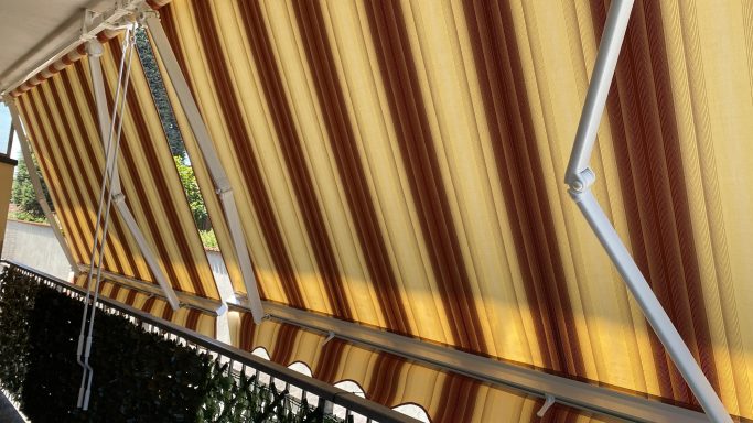 Tende da sole per balcone Striped yellow and brown awning partially extended over a balcony railing.