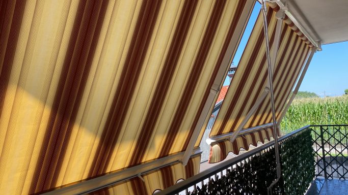 Tenda da sole per balcone Tende a righe giallo e marrone su un balcone con campo verde sullo sfondo.